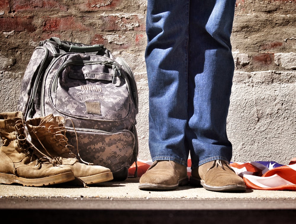 A minimalist picture depicting a US war veteran standing with his backpack, boots, and a US flag