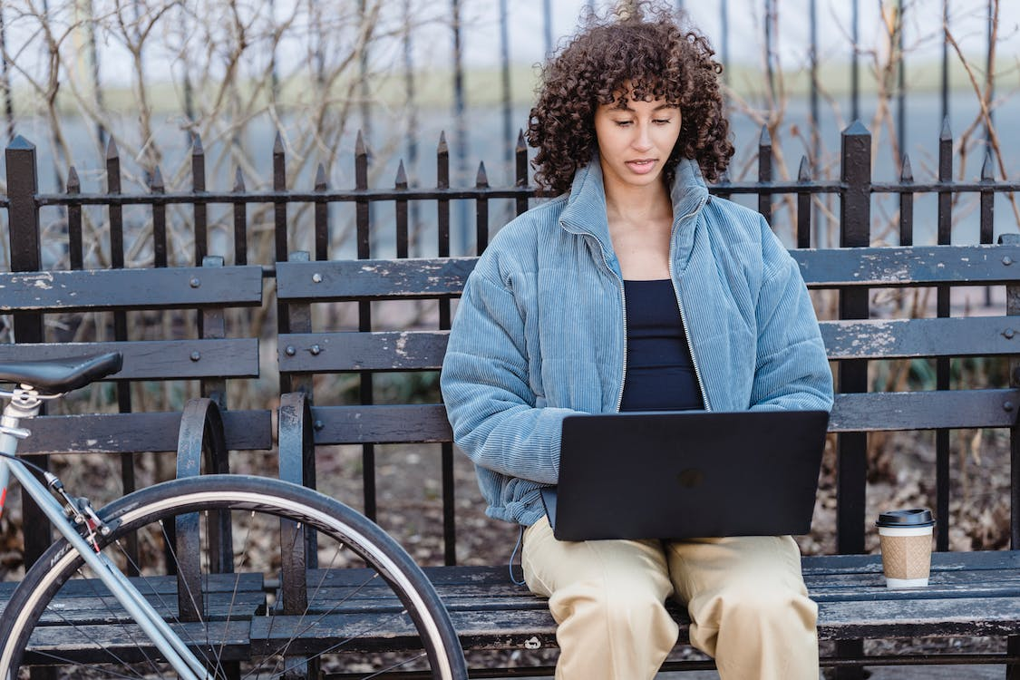 An image of a concentrated woman using a laptop while sitting on a bench in a park