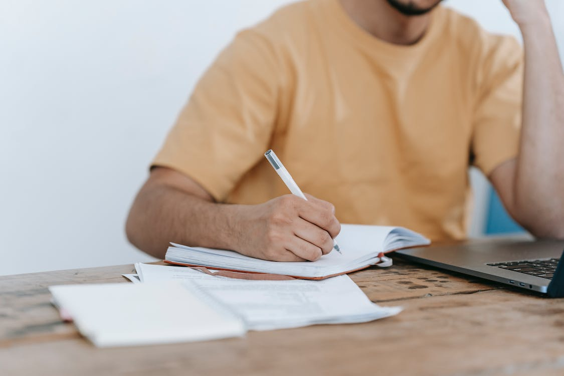 An image of a guy in yellow shirt writing on a notebook while sitting at a wooden desk in front of a laptop