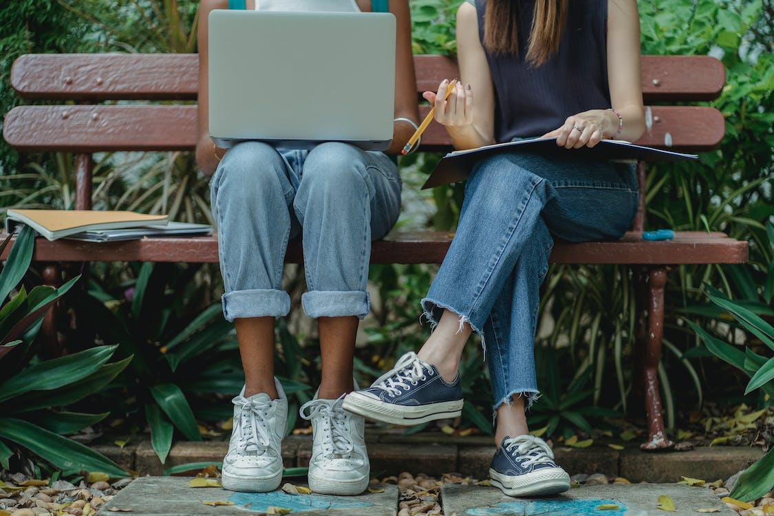 An image of a two female students wearing sneakers using their laptop while sitting on a bench in a park