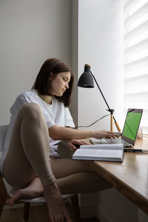 An image of a young woman reading a book while sitting at a wooden desk in front of a laptop